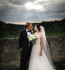 Groom and bride look at each other in wooded outdoor setting with sunlight peeking through clouds above them